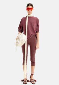 Bordowy bawełniany sweatshirt z kieszenią z przodu, zestawiony z pasującymi legginsami. Modelka trzyma w ręku paskowaną torebkę, nosi czerwone okulary przeciwsłoneczne i sandały z frędzlami.
