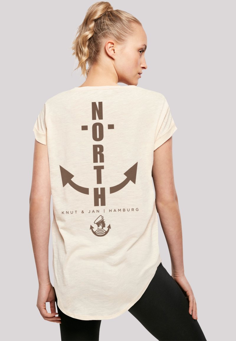 Cream-colored short-sleeve t-shirt featuring brown "NORTH" text and arrow graphics on the back, with a curved hem and soft texture.