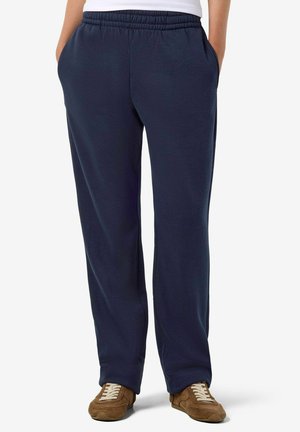 Personne portant un pantalon de survêtement bleu marine et des baskets marron, debout les mains dans les poches devant un fond blanc.