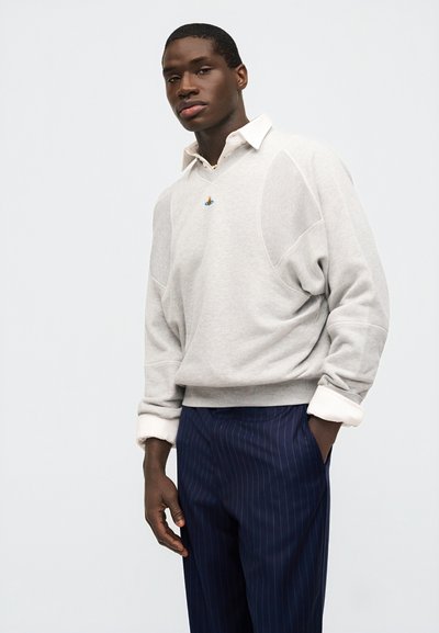 Άνδρας που φοράει ανοιχτό γκρι πουλόβερ με λογότυπο πάνω από λευκό πουκάμισο με γιακά και ναυτικό μπλε παντελόνι με ρίγες, ποζάροντας μπροστά σε απλό φόντο.