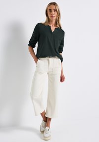 Donna con capelli biondi che indossa una maglietta henley a maniche lunghe scura, pantaloni cropped a gamba larga di colore bianco sporco e sneakers bianche, in piedi contro un muro bianco.