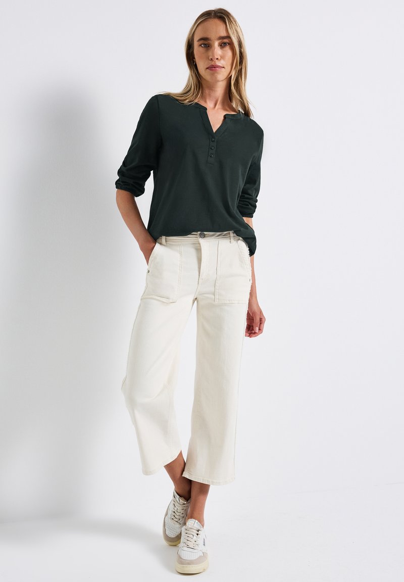 Donna con capelli biondi che indossa una maglietta henley a maniche lunghe scura, pantaloni cropped a gamba larga di colore bianco sporco e sneakers bianche, in piedi contro un muro bianco.