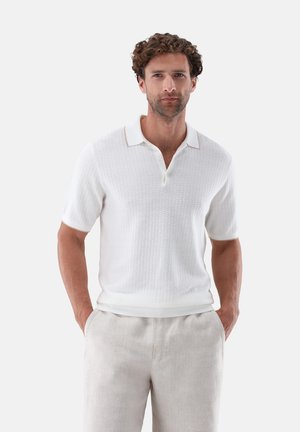 Mann mit lockigem Haar, der ein weißes, strukturiertes Polohemd mit kurzen Ärmeln und hellbeige Hose trägt, steht mit den Händen in den Taschen.