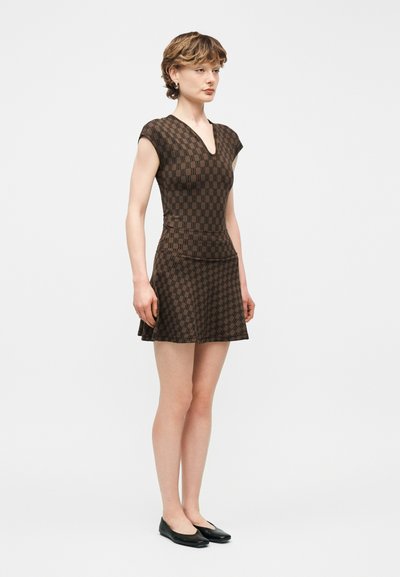 Νέα γυναίκα που στέκεται, φορώντας ένα καφέ φόρεμα με μοτίβα και κοντά μανίκια σε σχήμα V και μαύρες φλατ παπούτσια, σε απλό λευκό φόντο.