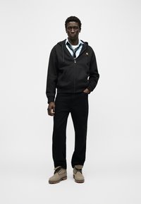 Νεαρός άνδρας στέκεται με το ένα χέρι στην τσέπη, φοράει μαύρο φούτερ με φερμουάρ, μπλε πουκάμισο με χαλαρή γραβάτα, μαύρο παντελόνι και μπεζ μπότες σε απλό φόντο.