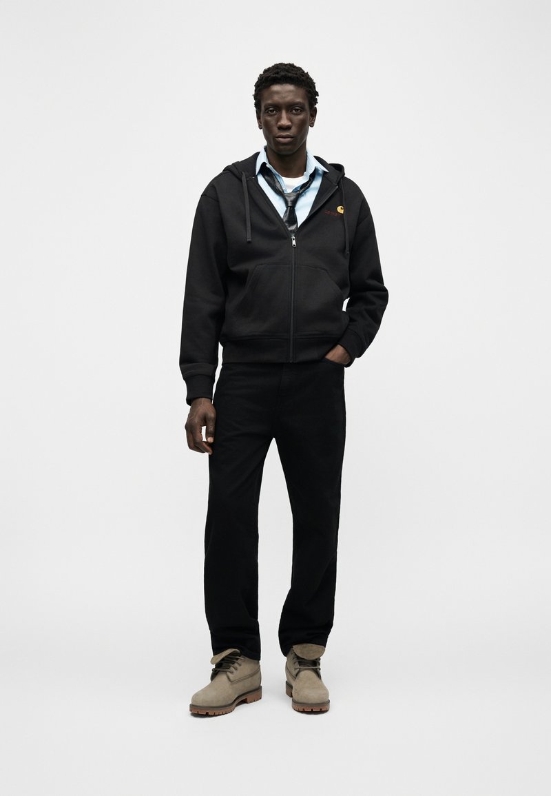 Junger Mann steht mit einer Hand in der Tasche, trägt eine schwarze Kapuzenjacke mit Reißverschluss, blaues Hemd mit locker gebundener Krawatte, schwarze Hose und beige Stiefel vor schlichtem Hintergrund.