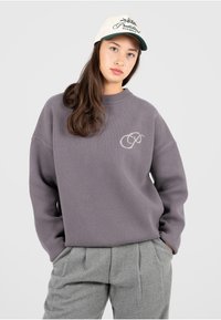 Pull en tricot violet avec une finition texturée et un col rond, comportant un logo brodé de couleur claire sur la poitrine.
