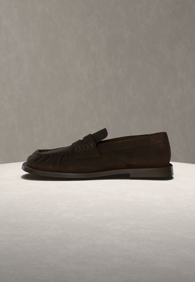 Σκούρο καφέ σουέντ παπούτσι τύπου penny loafer με επίπεδη σόλα, λεπτομέρειες ραφών και λουράκι στο επάνω μέρος, παρουσιασμένο σε φωτεινή επιφάνεια.