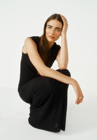 Robe noire texturée avec un haut ajusté et une jupe fluide, présentant un motif ondulé et nervuré. Le modèle est posé en position accroupie, la main sur la tête.