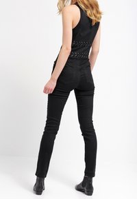 Top negro sin mangas con diseño de corsé que presenta detalles de encaje en la parte posterior, combinado con jeans negros ajustados y botas de tobillo.