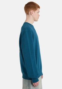 Φούτερ από βαμβάκι σε αποχρώσεις του πετρόλ, με κλασική στρογγυλή λαιμόκοψη και ριμπ τελειώματα στα μανίκια. Η πλάγια όψη δείχνει άνετη εφαρμογή και μινιμαλιστικό σχέδιο.