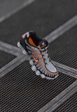 Gray and orange athletic shoe with distinct cushioning sole placed on a metal grid surface.