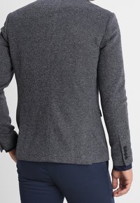 Uomo che indossa un blazer grigio a spina di pesce aderente e pantaloni blu navy, mostrato di spalle su uno sfondo neutro.