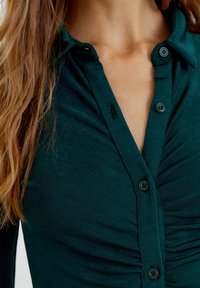 Chemise boutonnée vert foncé avec un col, présentant une texture froncée et des boutons noirs, vue rapprochée de la zone de la poitrine.