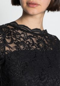 Černý krajkový top s propracovanými květinovými vzory a flitry, se vlnitým výstřihem a texturovanou černou látkou pod ním.