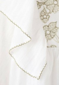 Bílá textilie s texturovaným vzorem. Má obloukový okraj se zeleným stehem a krajkovými doplňky, zdobená květinovou výšivkou.