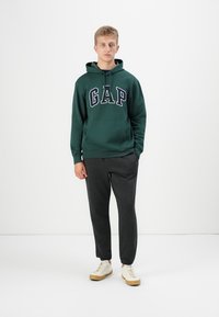 Green hoodie with a front pocket and blue "GAP" lettering, paired with dark gray sweatpants and beige sneakers.