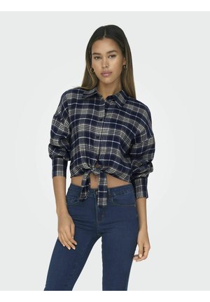 Camicetta cropped a quadretti blu navy con maniche lunghe, chiusura a bottoni e orlo annodato; abbinata a jeans aderenti blu scuro.