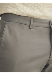 Lähikuva beigestä neuleesta, joka on tukkitettu korkeavyötäröisiin harmaisiin housuihin, joissa on vyölenkit ja sivuvetoketjukiinnitys.