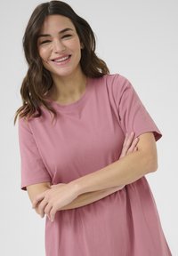Krótki różowy t-shirt z okrągłym dekoltem i krótkimi rękawami, charakteryzujący się luźnym krojem i gładką fakturą. Model uśmiechający się, z założonymi rękami.