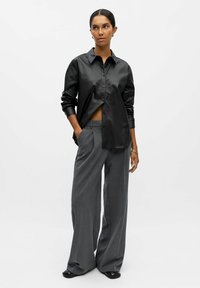 Zwarte faux leren shirt met een knopsluiting, lange mouwen en een losse fit, gecombineerd met grijze wijde broek en zwarte schoenen.