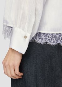 Biała plisowana bluzka z koronkowym wykończeniem na dole, z zapinanym mankietem z detalem w postaci perły, oparta na ciemnym dżinsowym materiale.