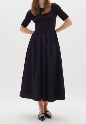 Frau trägt ein schwarzes Midikleid mit kurzen Ärmeln und einem gerafften Rock, kombiniert mit schwarzen spitzen Ballerinas mit Riemen.