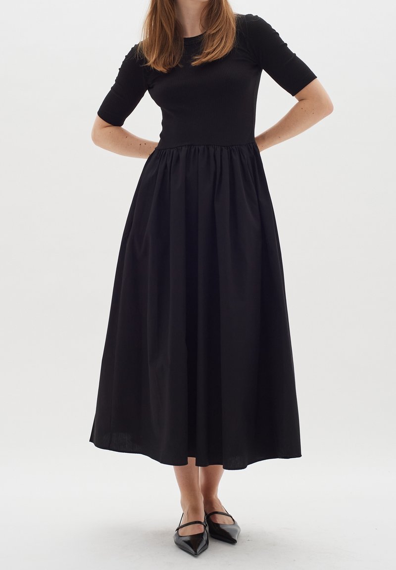 Femme portant une robe midi noire à manches courtes et jupe froncée, associée à des ballerines noires pointues avec des brides.