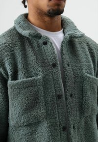 Groene fleecejas met een gestructureerde, pluizige afwerking. Voorzien van een kraag, knoopsluiting en twee voorzakken.