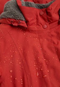 Giacca impermeabile rossa con un morbido colletto in pile grigio, caratterizzata da gocce d'acqua sulla superficie e bottoni a pressione lungo la parte anteriore.
