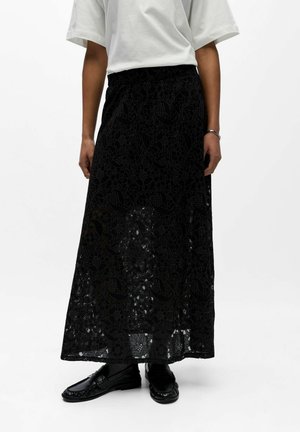 Kobieta ubrana w długą czarną spódnicę z kwiatowej koronki, biały krótki rękaw, czarne mokasyny i srebrną bransoletkę na prawej ręce.