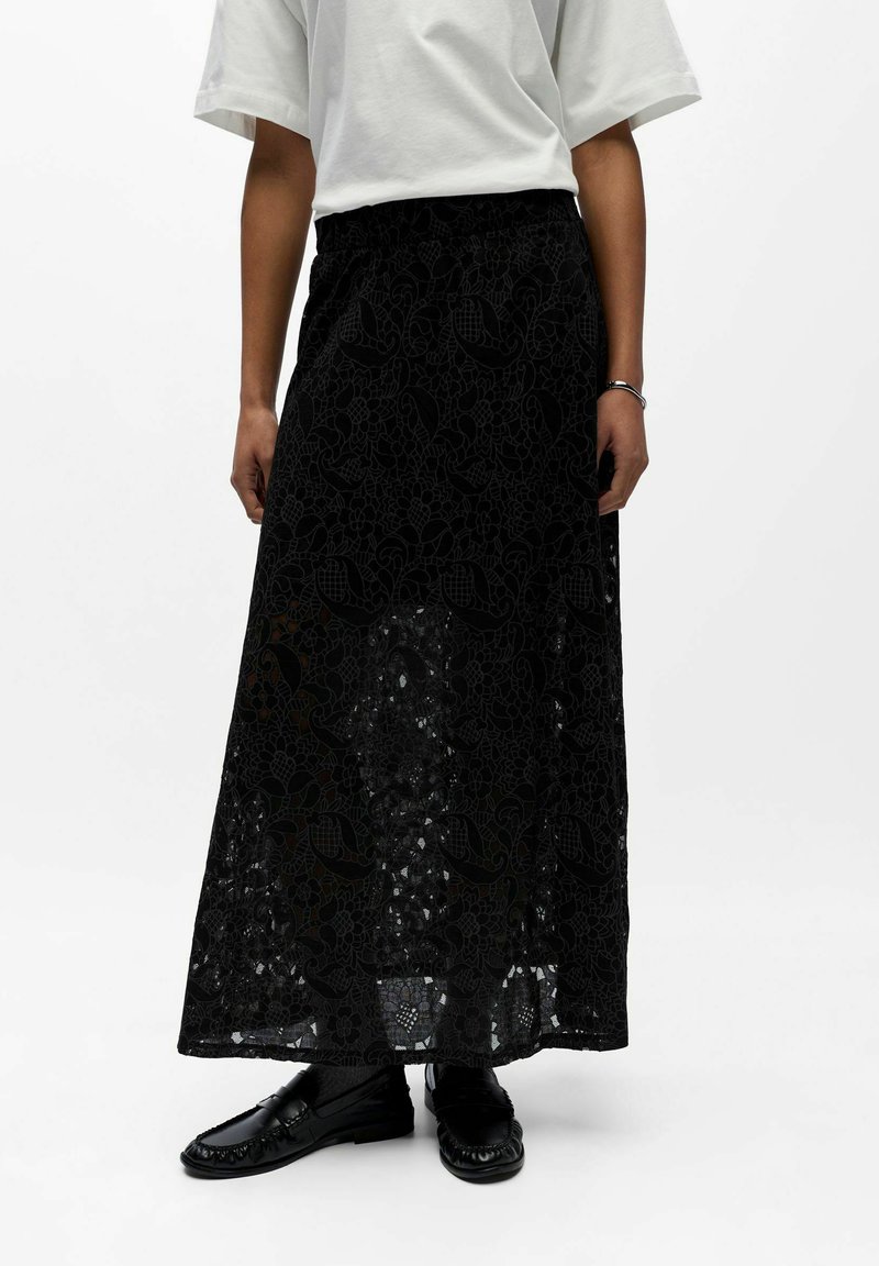 Femme portant une longue jupe florale en dentelle noire, un haut blanc à manches courtes, des mocassins noirs et un bracelet argenté au poignet droit.