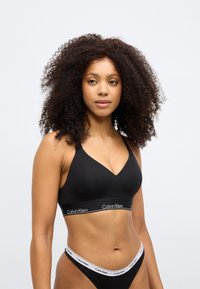 Crna Calvin Klein braleta s V-izrezom i širokom osnovom s bijelim logotipom. Tkanina je glatka i rastezljiva. Jednostavan i minimalistički dizajn.