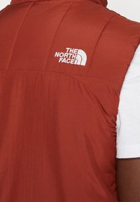 Chaleco acolchado rojo con diseño de cuadrícula, con un logo bordado blanco en el hombro izquierdo. El material parece suave y ligero.