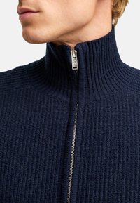 Granatowa, prążkowana, dzianinowa kurtka zapinana na zamek z wysokim kołnierzem i srebrnym detalem zamka. Miękka textura z wąskimi pionowymi wzorami.