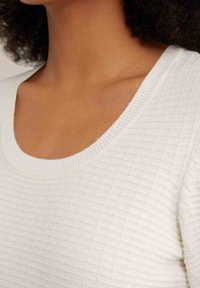 Bílý texturovaný svetr s kulatým výstřihem, žebrovanými manžetami a jemným kostkovaným vzorem, zaměřující se na měkkost a pohodlí.