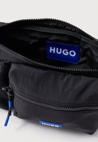 Μαύρη τσάντα με φερμουάρ από υφασμάτινη υφή. Διαθέτει μια μπλε ετικέτα με το "HUGO" και μια τσέπη με φερμουάρ με μπλε γάντζο. Ο εσωτερικός χώρος είναι επενδυμένος με μαύρο υλικό.