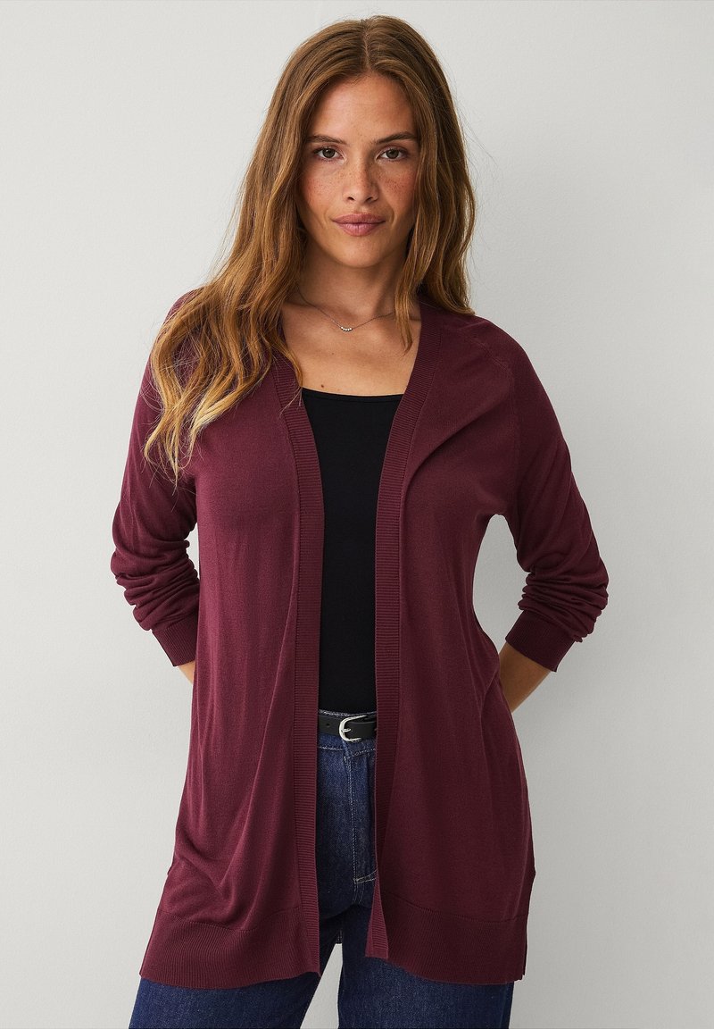 Bordeaux lichte cardigan met lange mouwen, open voorkant en geribbelde manchetten. Gecombineerd met een zwarte top en blauwe jeans. Zachte textuur.