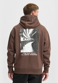 Un sweat à capuche marron avec un grand motif noir et blanc au dos, représentant une plateforme de train souterraine courbée avec du texte en dessous.