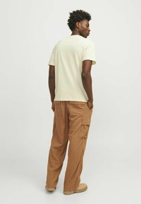 Camiseta de algodón color beige claro, de cuello redondo, combinada con pantalones cargo marrones con bolsillos laterales, corte relajado y detalles de costura visibles.