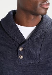 Man som bär en marinblå stickad tröja med sjalkrage och två knappar över en vit crewneck-skjorta.