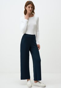 Witte lange mouw top gecombineerd met marineblauwe wijde broek met ribstructuur en zijslits, afgewerkt met witte sneakers.