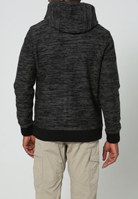 Man i en mörkgrå mönstrad hoodie och beige cargobyxor, sedd bakifrån mot en enkel bakgrund.