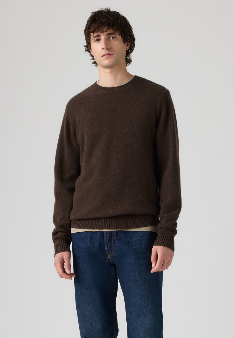Jeune homme portant un pull en maille marron foncé sur une chemise beige et un jean bleu, debout devant un fond clair uni.