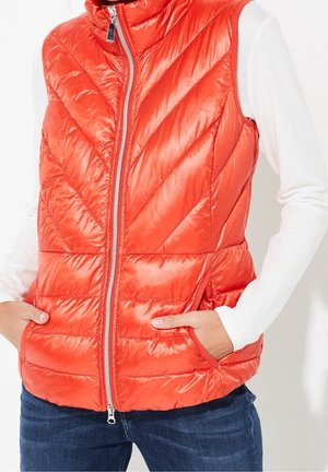 Polstret orange ærmeløs vest med frontlynlås og lommer, båret over en hvid langærmet skjorte og blå jeans.