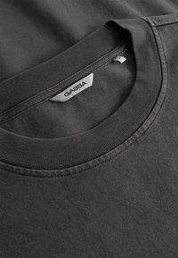 Približen posnetek temno sive majice z zaobljenim izrezom in belo etiketo znamke "GABBA" všito na notranji strani ovratnika.