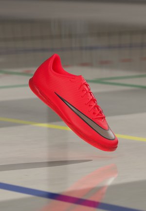 Chaussure de sport rouge vif avec un swoosh Nike noir flottant au-dessus d'un terrain intérieur avec des lignes colorées.