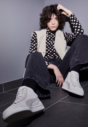 Jeune personne aux cheveux bouclés assise par terre, portant des baskets blanches montantes, un jean noir, une chemise à motifs de cœurs et un gilet clair contre un mur gris.