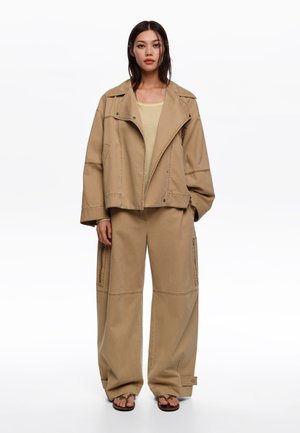 Mujer joven de pie con una chaqueta holgada color marrón claro, pantalones anchos a juego con bolsillos cargo, camiseta beige clara y sandalias de punta abierta.