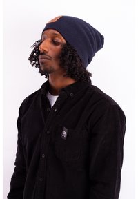 Navy knitted beanie with a leather patch, worn with a black corduroy button-up shirt featuring a pocket and a small brand logo.